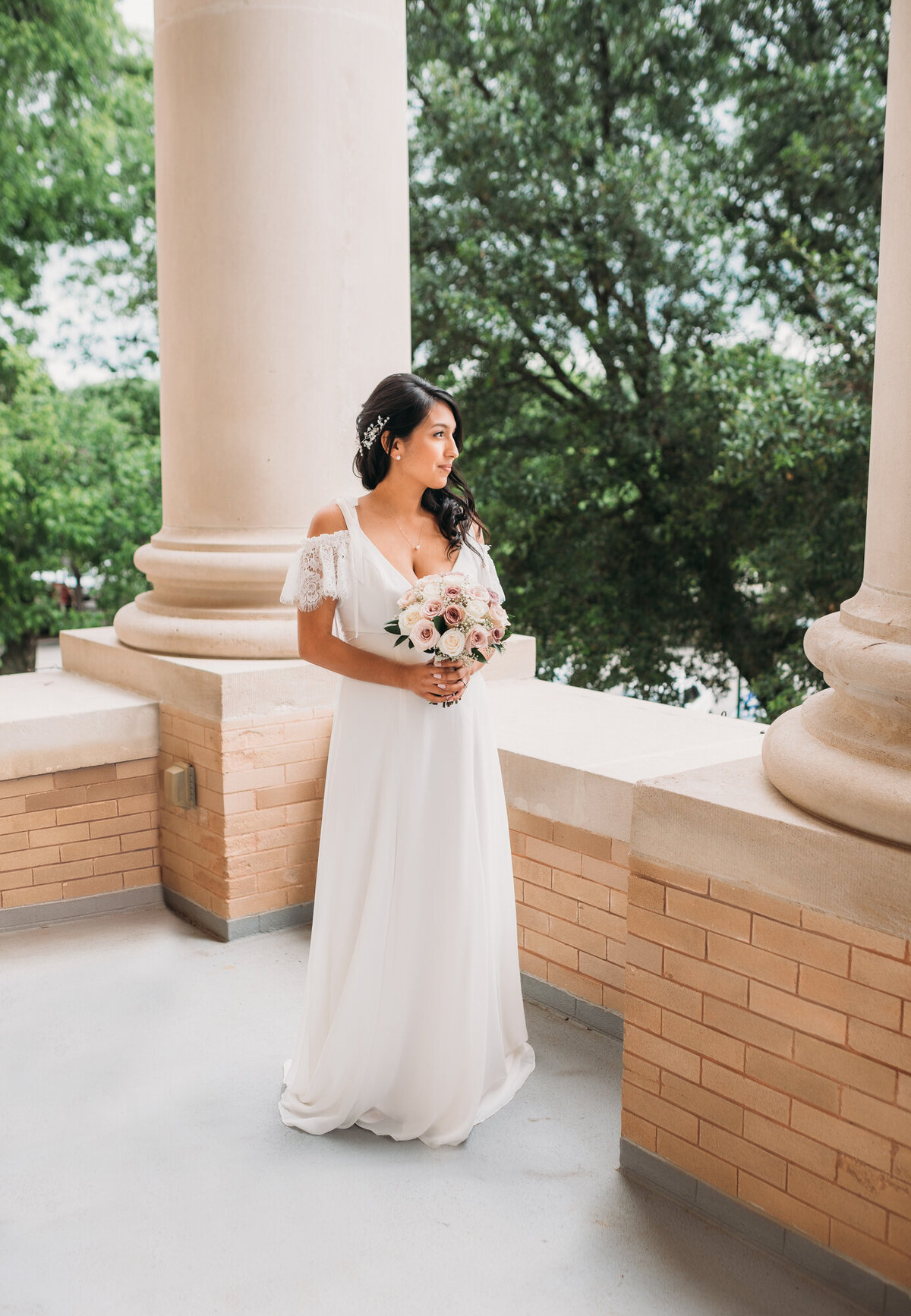 Couples Photography, Woman in a wedding dress stands on a patio and looks off into the distance holding a small flower bouquet.