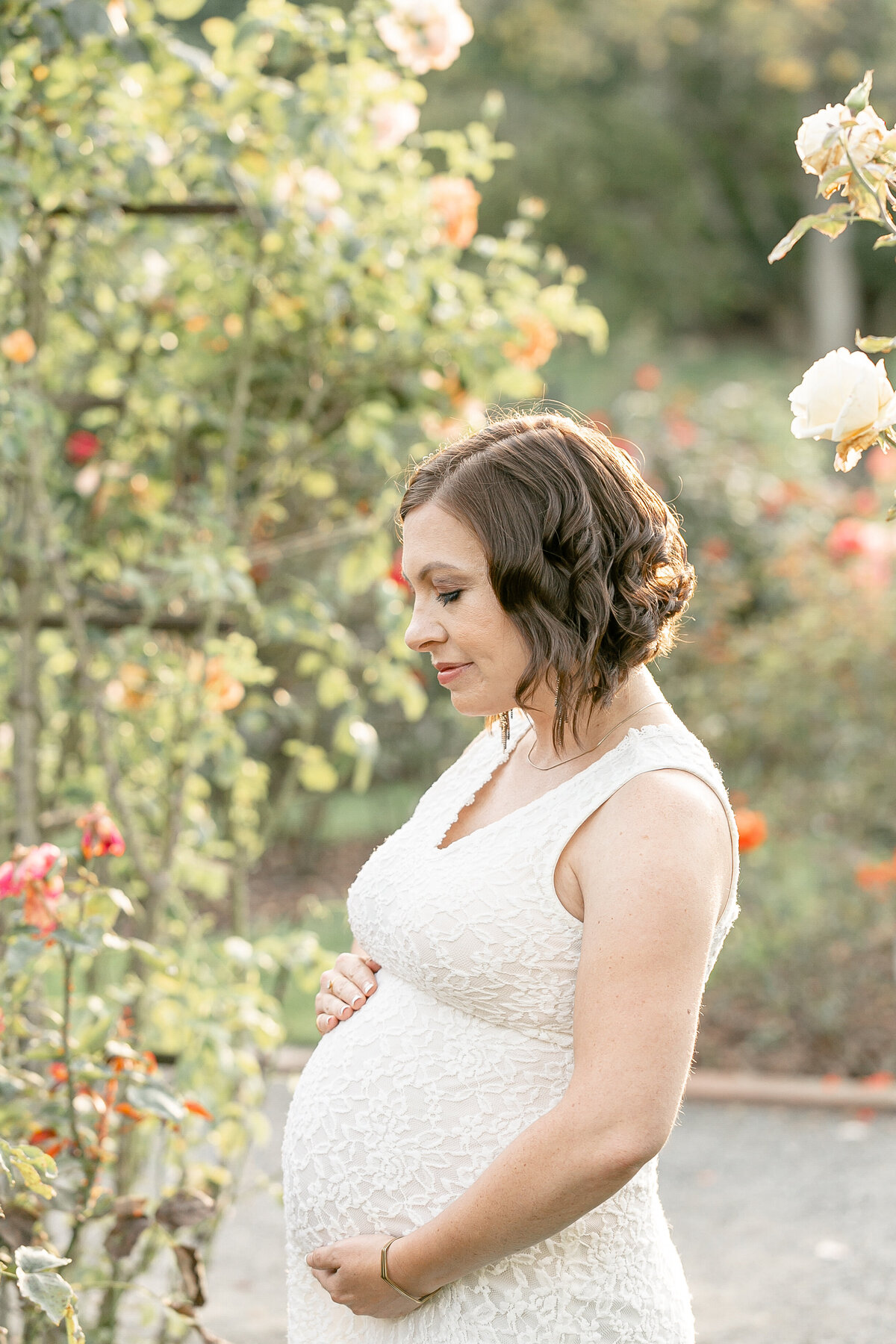 Pregnant Woman dressed in white lace sleeveless dress standing in Portland Rose Garden. Photography Session with Portland Maternity Photographer