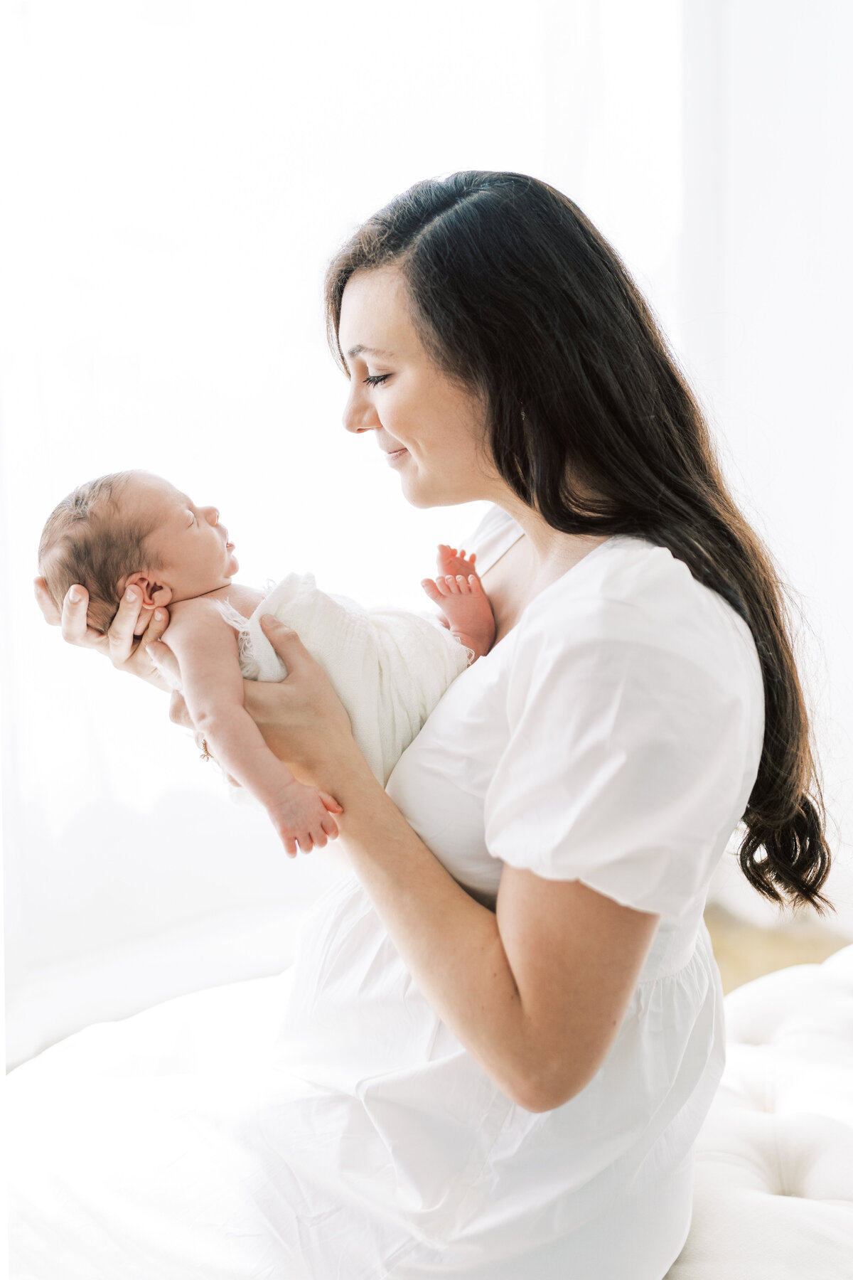 Portrait of a woman wearing a white dress and holding a swaddled newborn baby in front of her in a light and airy room.