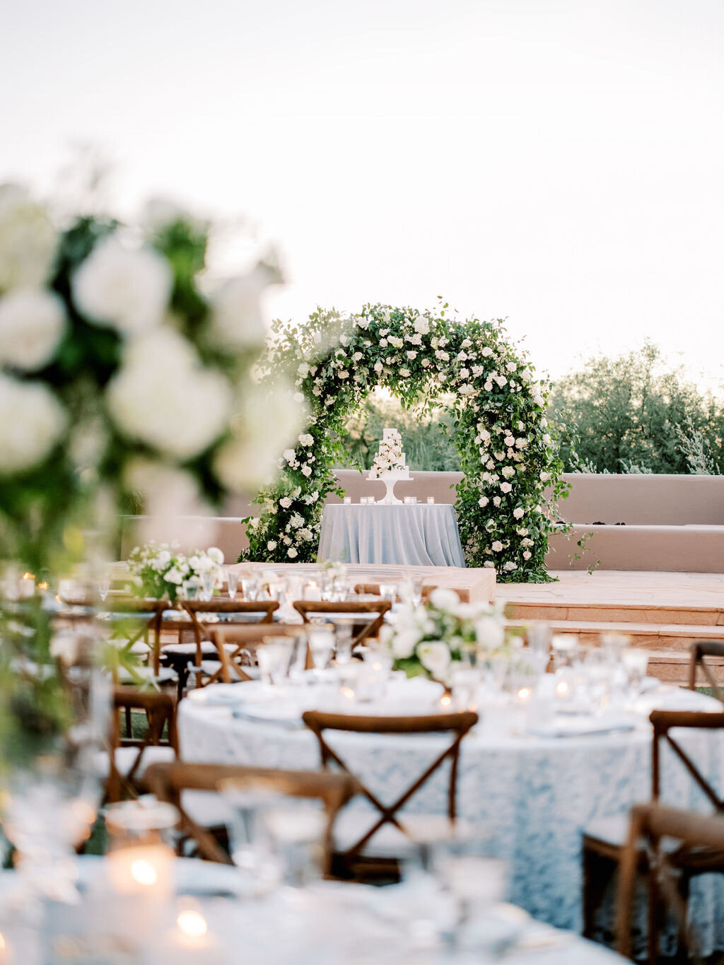 A breathtaking outdoor wedding reception featuring lush floral arches, a beautifully set dinner table, and a dreamy wedding cake display. This luxury wedding setup exudes timeless romance and sophistication, perfect for an elegant celebration.