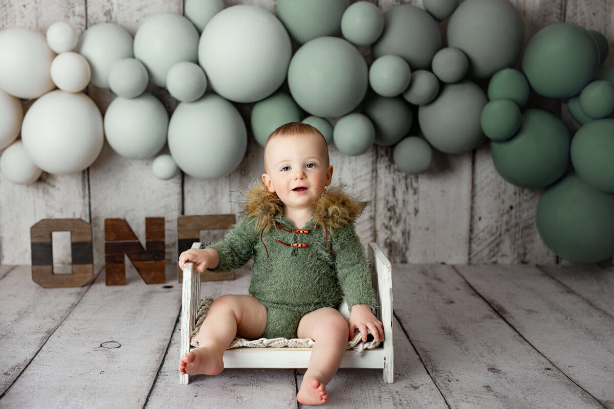 one year old boy in a green fuzzy outfit sitting on a white wooden bed with green balloons behind him and wooden one letters
