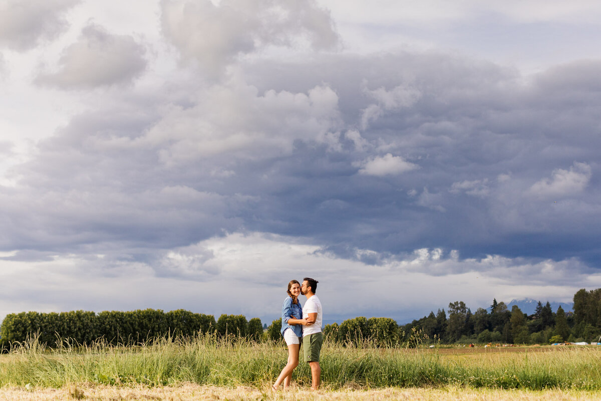 Engagement session in fields with moody stormy sky at Craven Farm wedding venue in Snohomish near Woodiville WA willow tree happy couple laughing colorful candid photo by Joanna Monger Photography