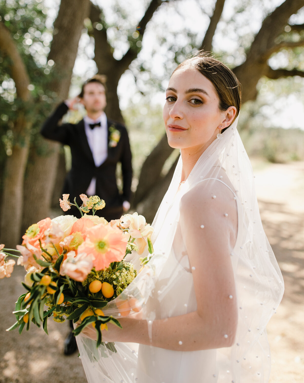 Bride with groom in background holding bouquet of bright orange and yellow florals