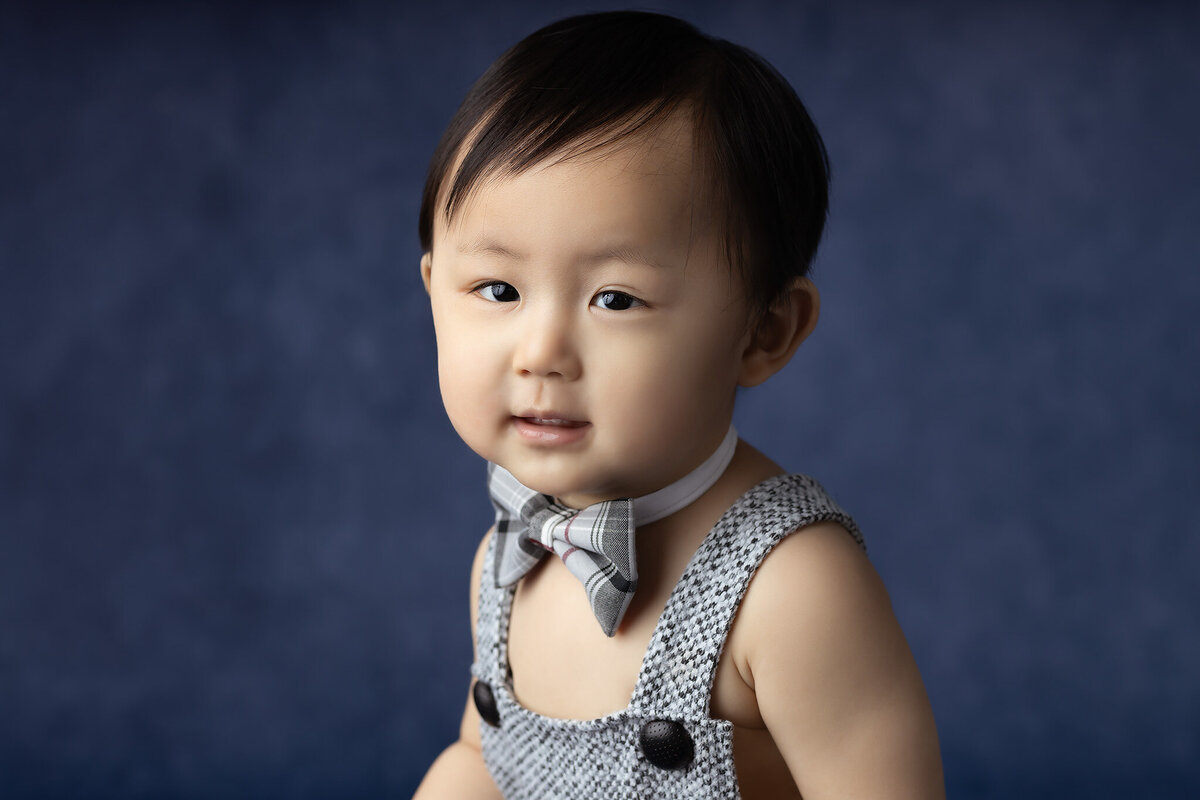 A baby with a bowtie smiles softly at the camera against a dark blue background.