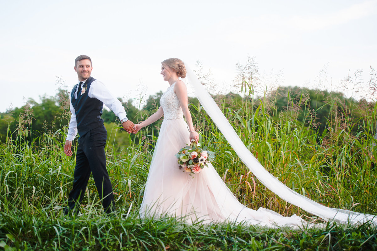 Newly married couple holds hands as they walk through tall grasses at the Chandler Hill Vineyard in Defiance, MO.