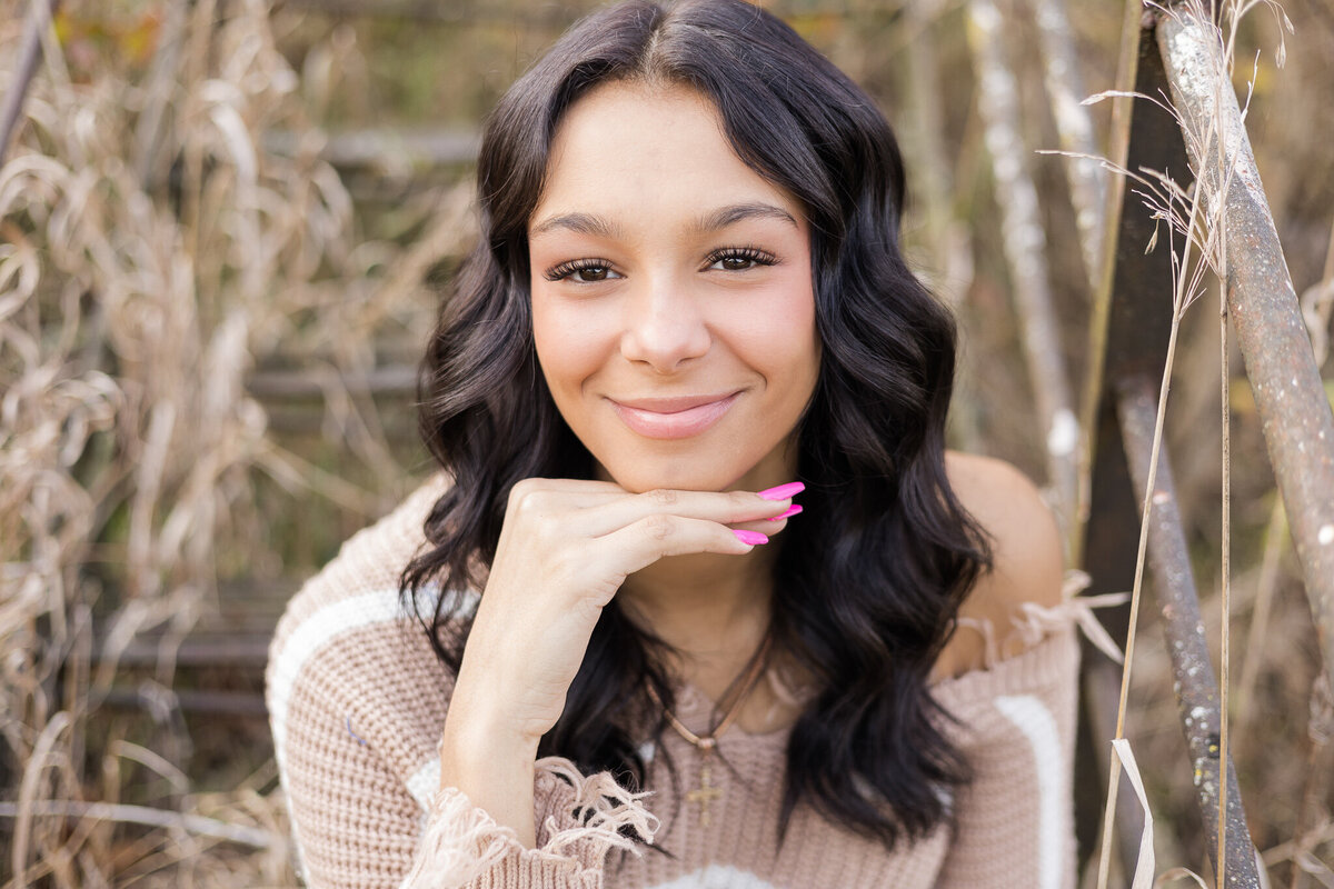high school female senior, with long black hair, sitting, looking at the camera smiling softly, wearing a light tan and white stripped shirt, with her hand under chin