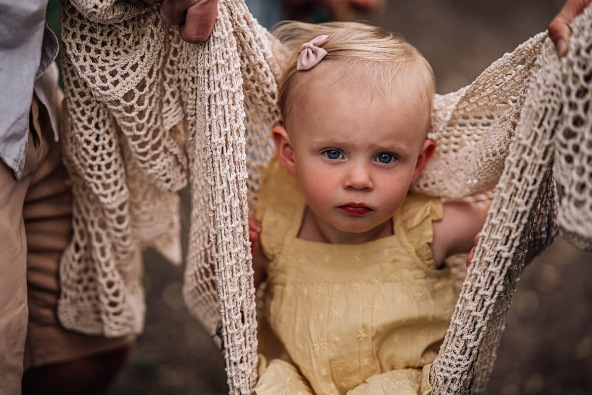 A toddler looks stoically into the camera while being held between her parents in an off white bohemian crocheted blanket.