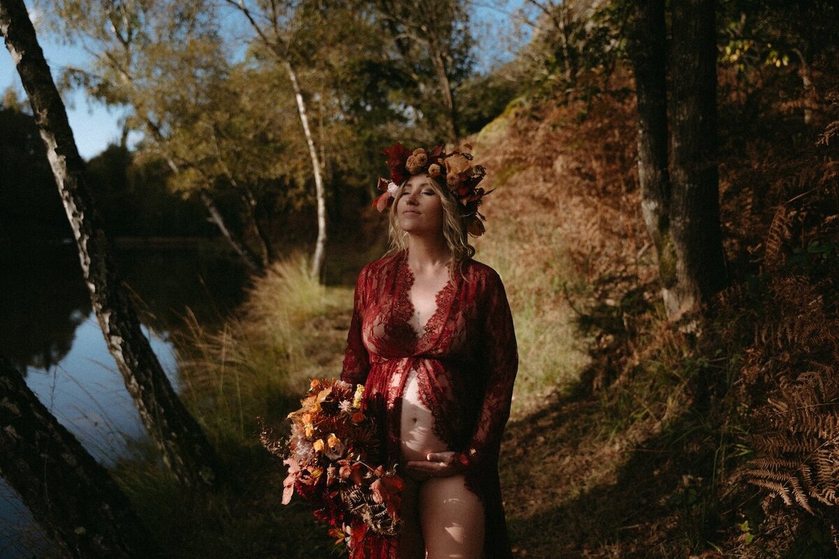 Femme enceinte en robe de dentelle rouge, bouquet automnal et couronne de fleurs sur la tête. Photographe Kamescop, séance grossesse en forêt bretonne.