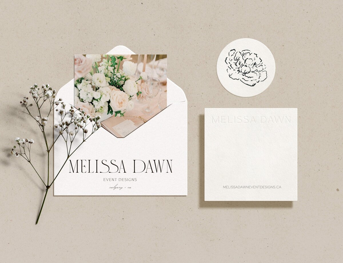 Invitation set including an envelope with a floral photo, a name card reading "Melissa Dawn Event Designs," a round floral sticker, and a small branch of white flowers on a textured background. Perfect for showcasing the elegance that brand identity design services can bring to any event.