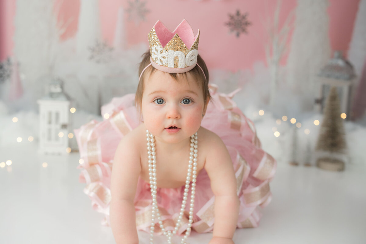 One year girl in crawling stance looking at camera wearing pearls, one year birthday crown and a pink tutu