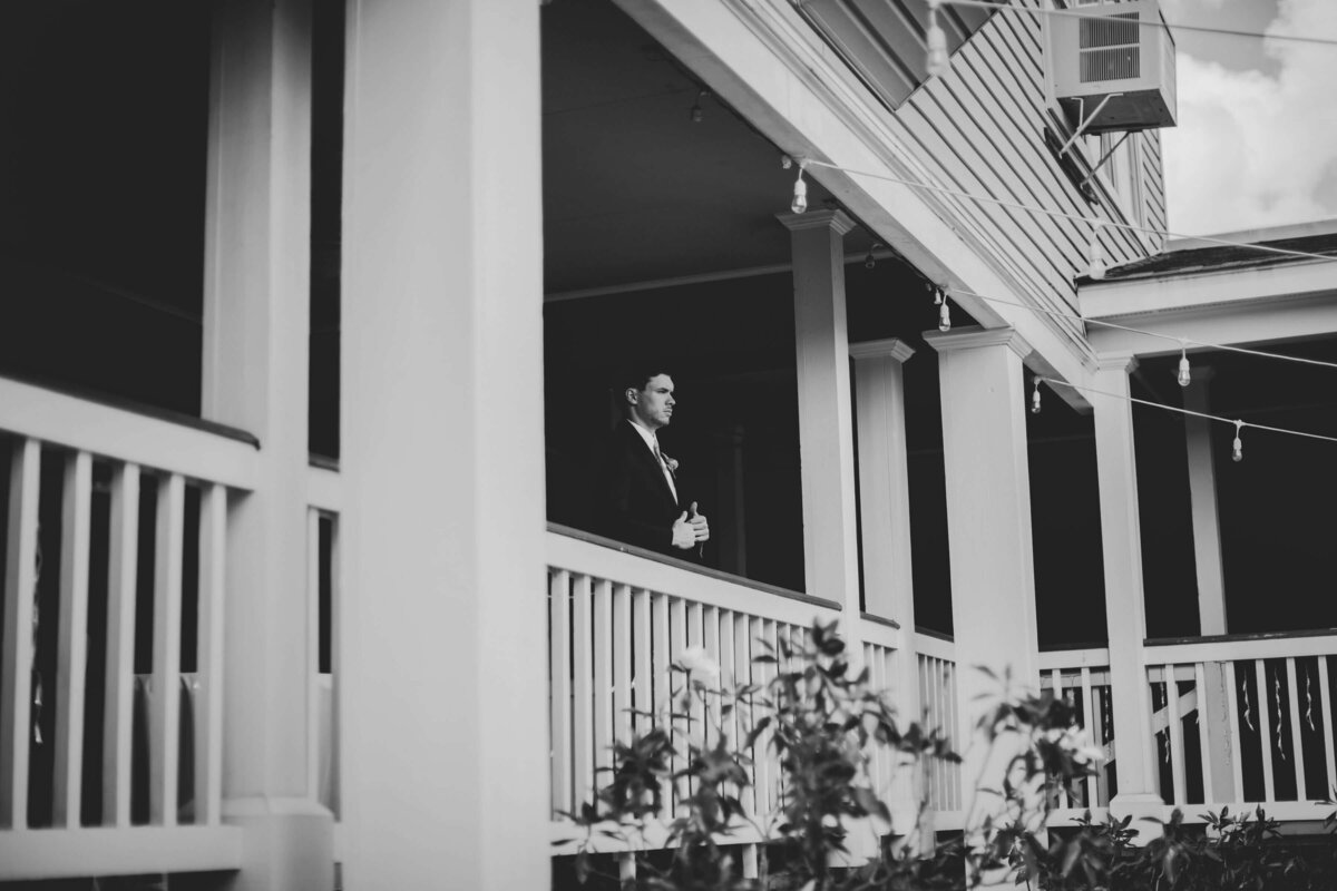 A man in a suit stands alone on a balcony, surrounded by columns and railings. The image is in black and white, with plants visible in the foreground.