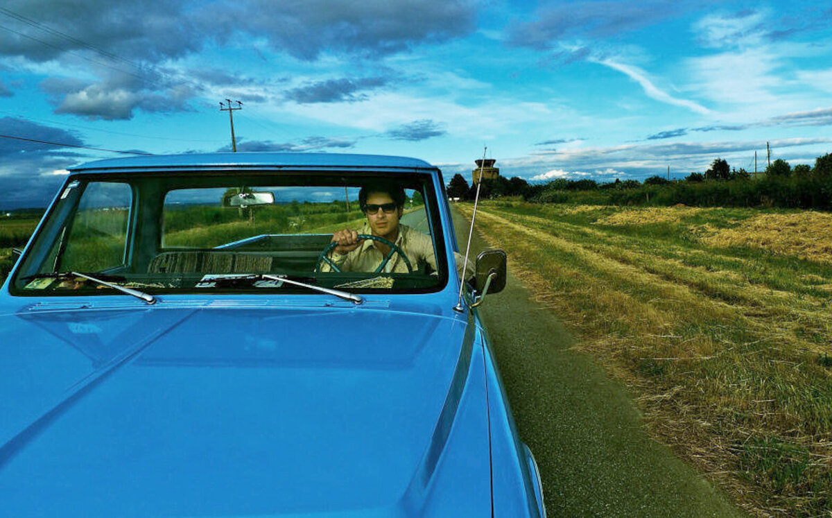 Musician Portrait Dustin Bentall driving vintage blue truck down country road wearing sunglasses blue sky with clouds behind him Roadshow Gallery