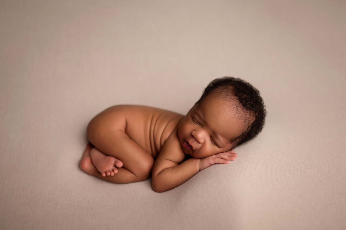 Newborn baby peacefully sleeping on a beige blanket, posed with hands under cheek during a professional portrait session in Waukesha, Wisconsin