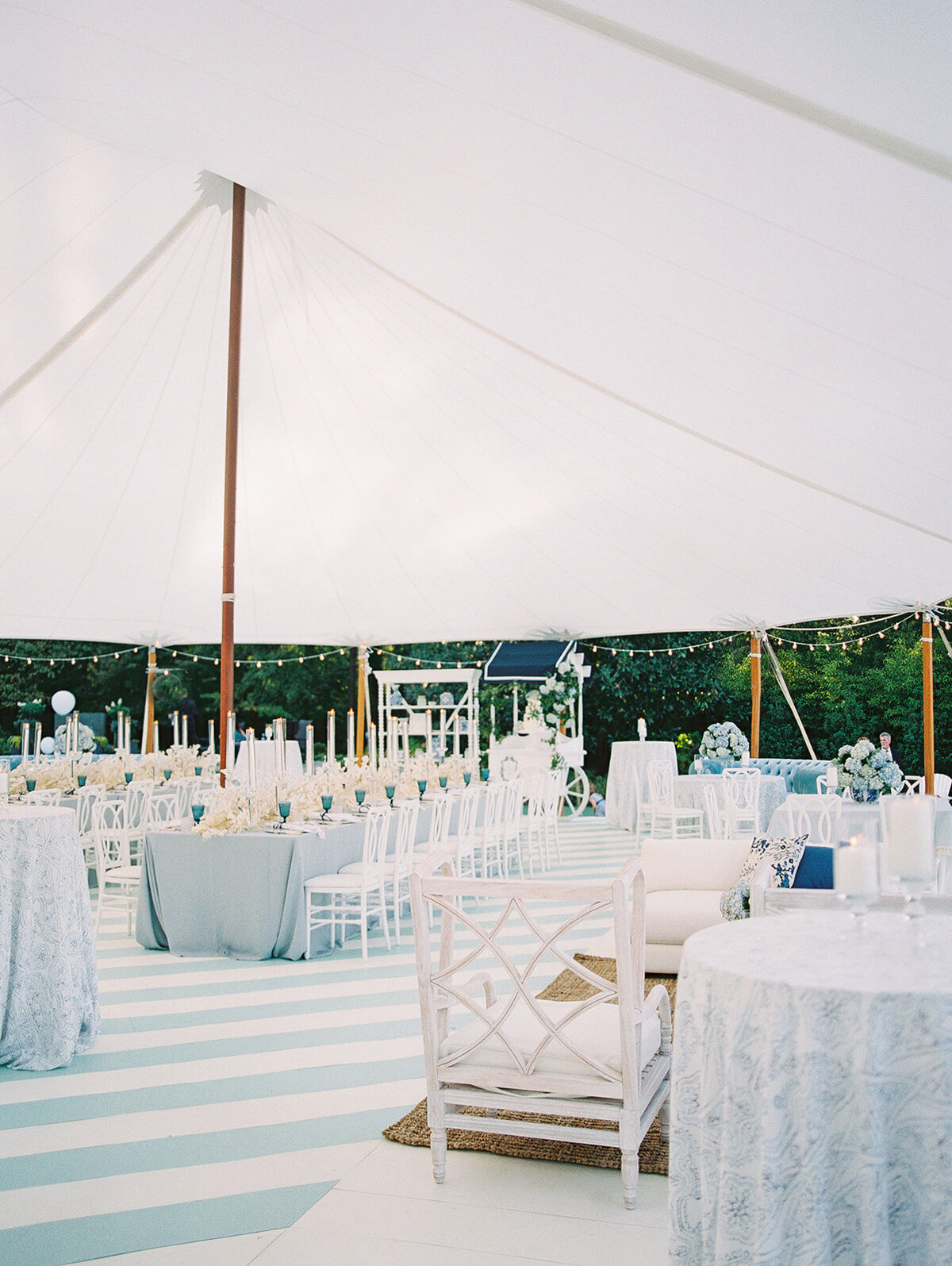 An elegant outdoor tented event setup with white decor, round tables with light blue tablecloths, and white chairs. The venue features a high, white canopy, floral arrangements, candles, and subtle lighting, creating a serene and sophisticated atmosphere.