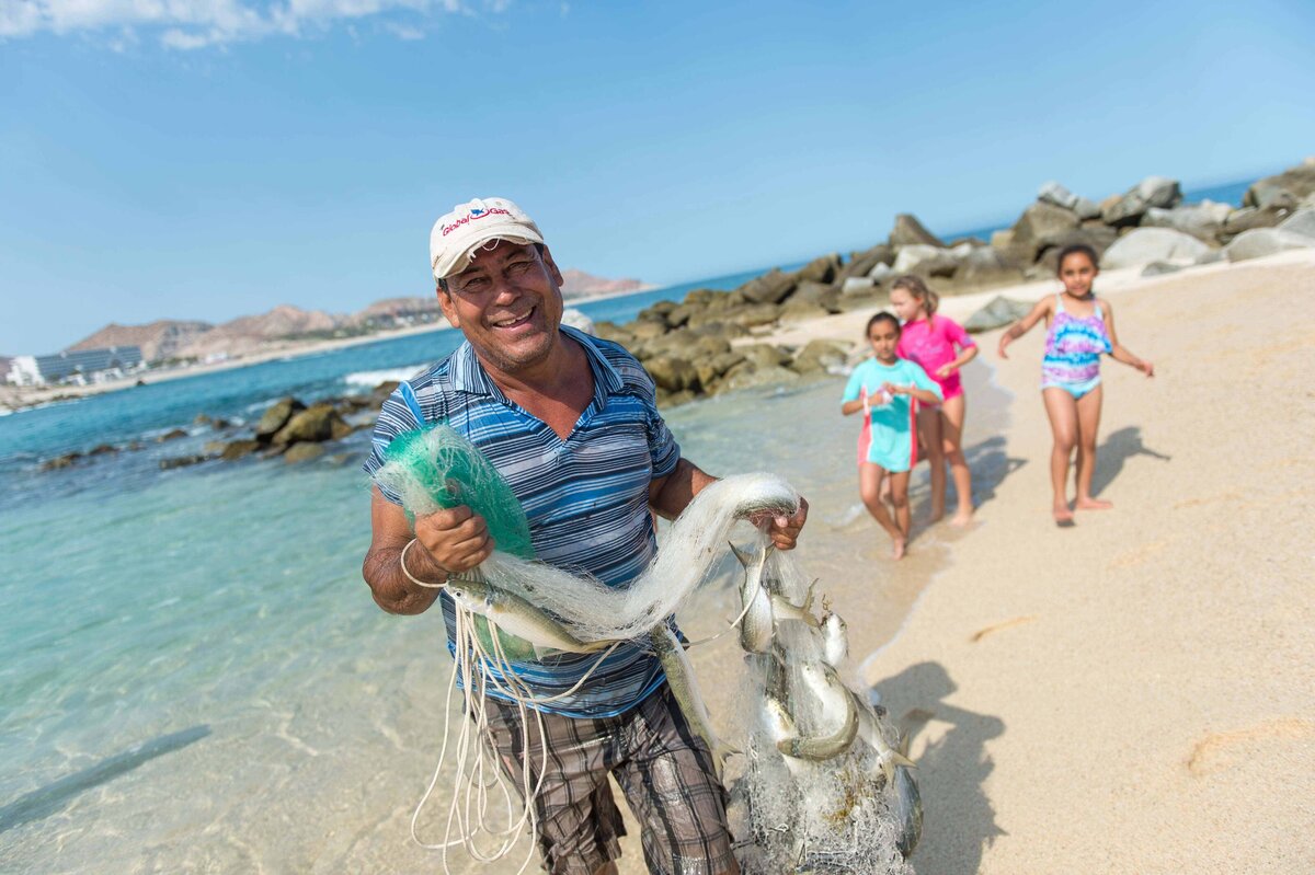 A fisherman in Baja smiles at camera and stands with a net full of fish in Baja Mexico outside the Hilton as children  run to see