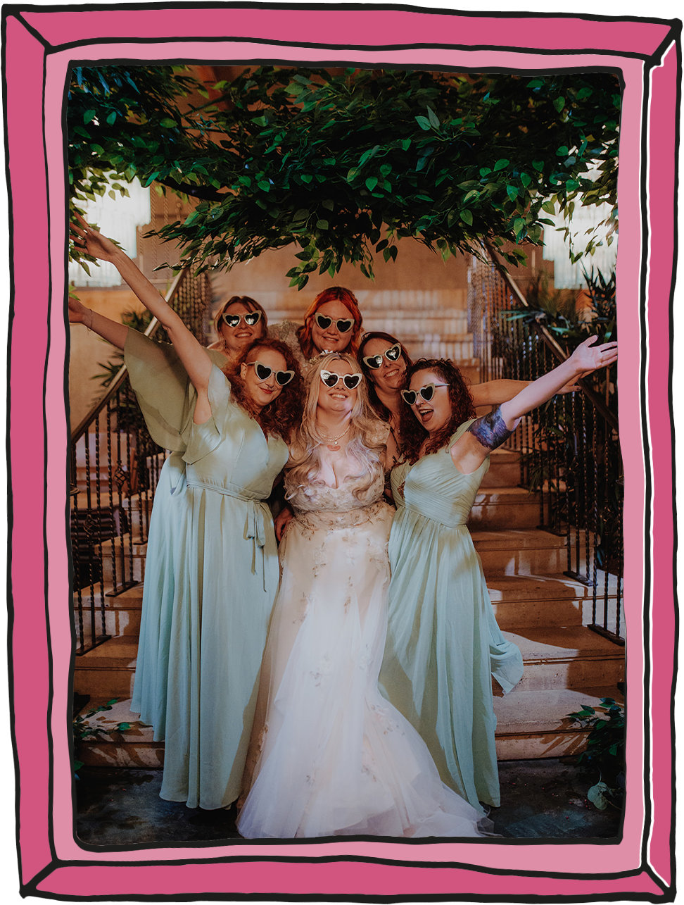 Bride in white wedding dress, surrounded by bridal party in pale green dresses with hands in the air. All wearing heart sun glasses