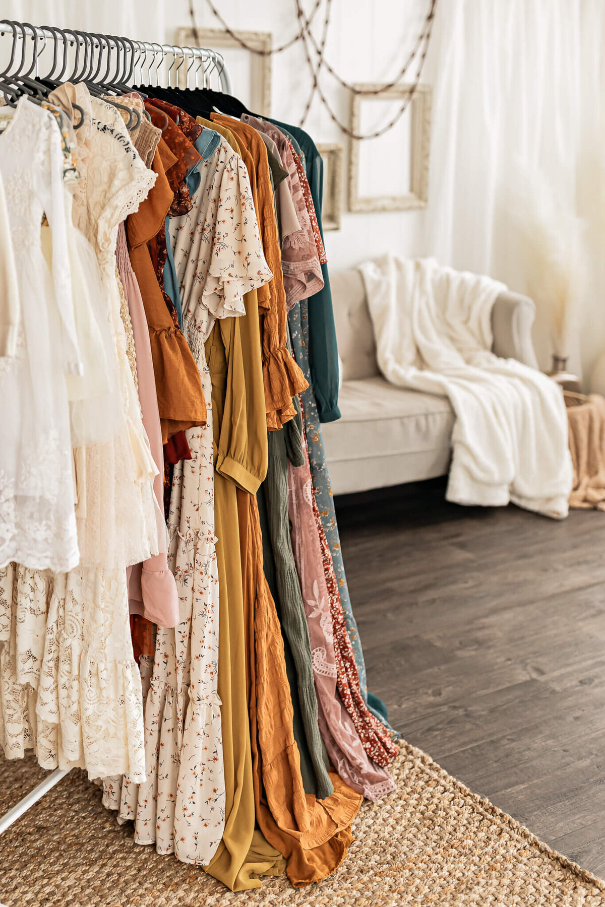 A clothing rack with an assortment of long, flowing dresses in various colors and patterns stands on a woven rug in a cozy room. A beige sofa draped with a white blanket is in the background, along with framed wall decor.