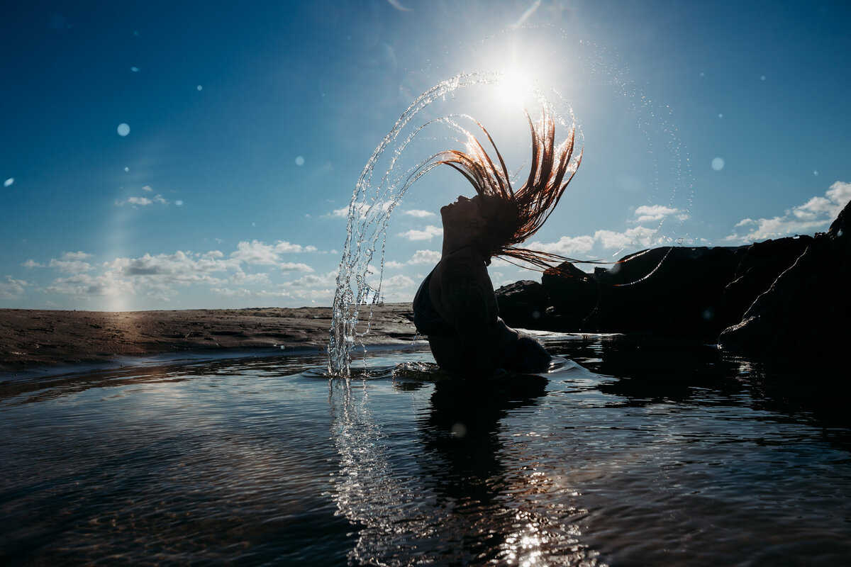 Woman flipping wet hair in a dramatic arc with water droplets catching sunlight during an empowering Salted Summer session at sunrise.