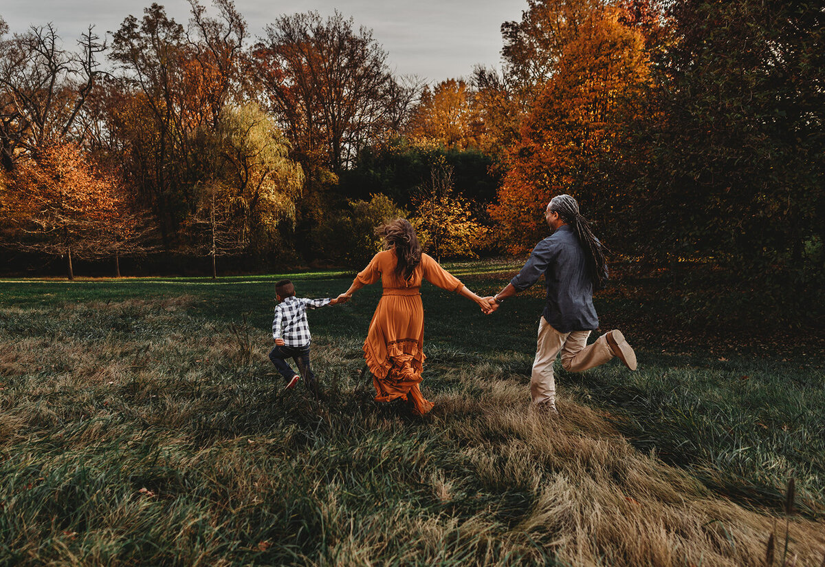 Family photographers Maryland photographs candid family pictures with mother, father and small child holding hands and running away from the camera to a tree line in a park for their fall family pictures