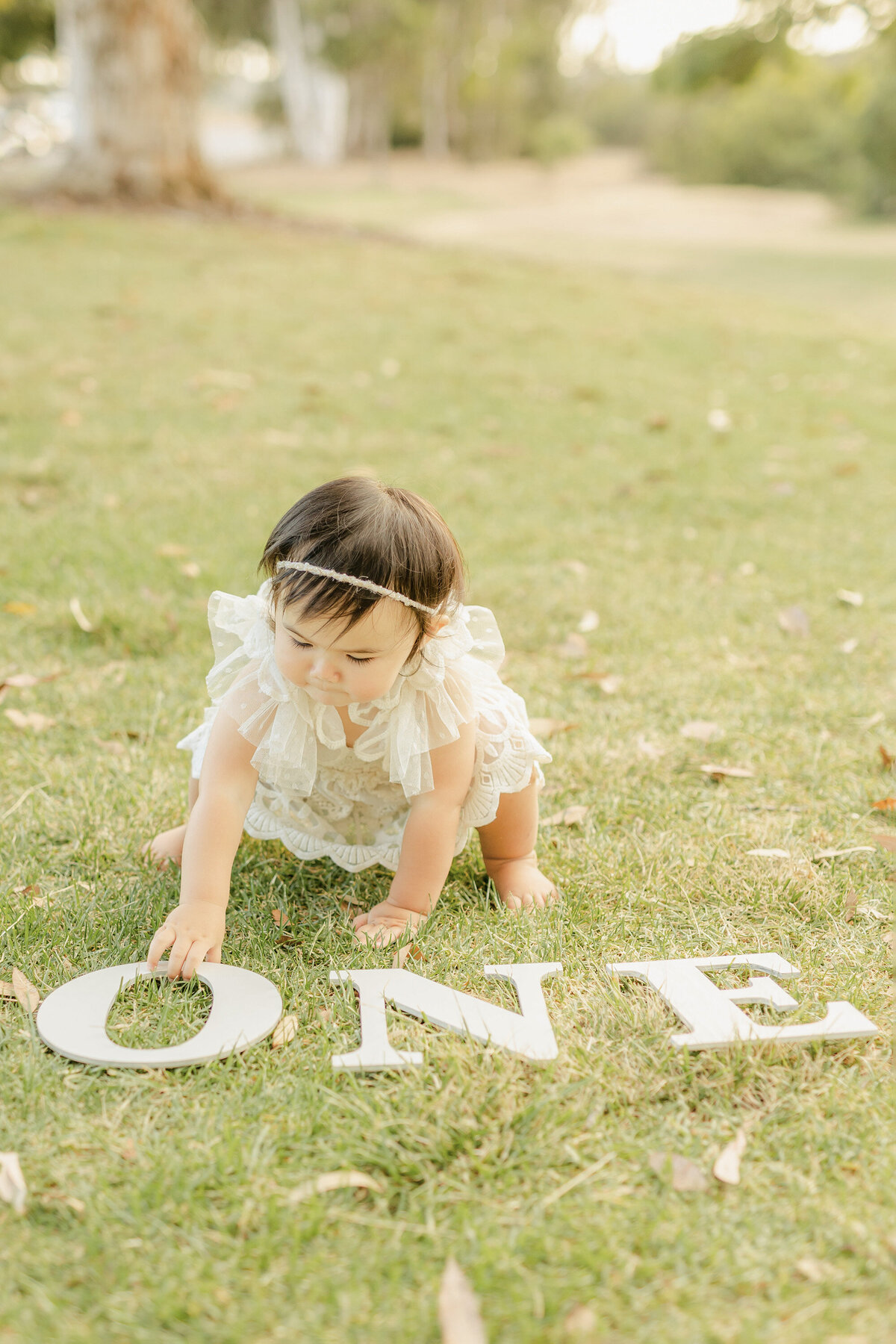 Adorable 1 year old baby girl crawling on the grass with white “ONE” letters in front of her during a San Diego milestone photoshoot. Captured by San Diego Family Photographer Yuka Schad.