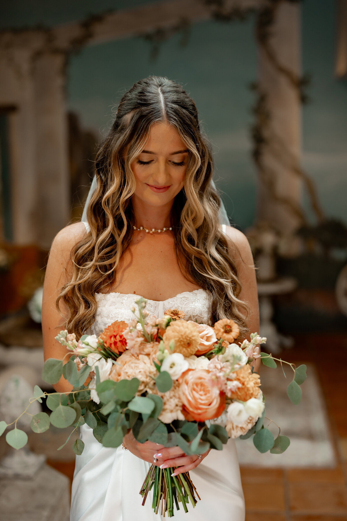 A bride in a white dress, captured by a New England wedding photographer, holds a bouquet with roses and greenery. She has long wavy hair and looks down with a slight smile, set against a backdrop of delicate, nature-inspired decor.