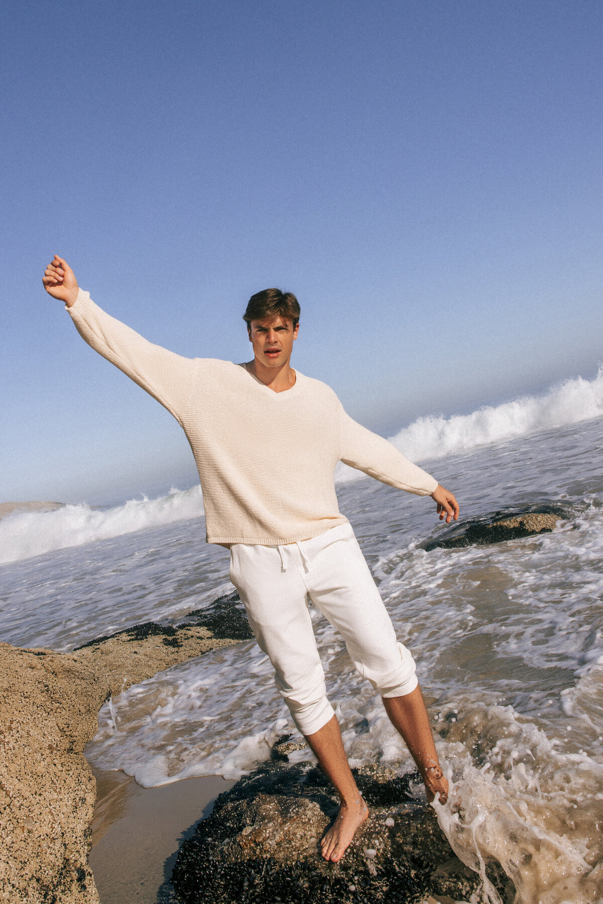 Male model in a cream sweater and rolled-up white pants balancing barefoot on coastal rocks as ocean waves crash around him in Cape Town. Captured mid-motion with arms outstretched against a clear blue sky.