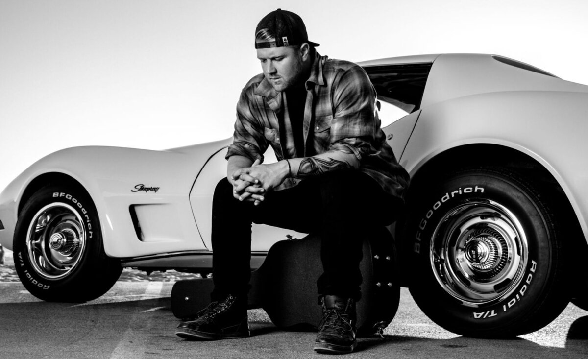 Male musician photo black and white Jake Parr wearing plaid shirt backwards baseball hat sitting upon black guitar case against vintage white Corvette