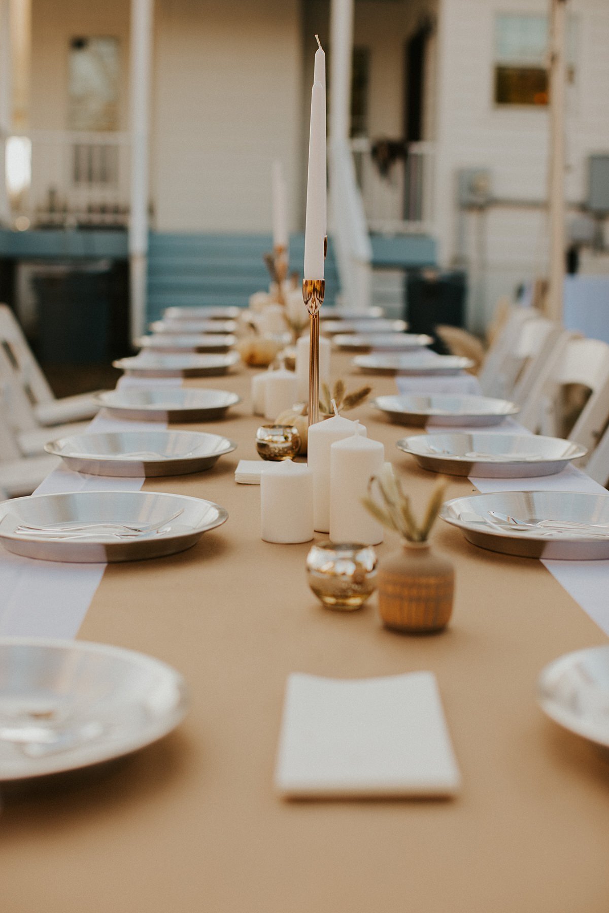 Long family style dinner table with white and gray china plates with a long tan table runner over a white table cloth. The table is decorated with white pillar candles, gold votive candles,  and tall gold candles sticks with white table candles.