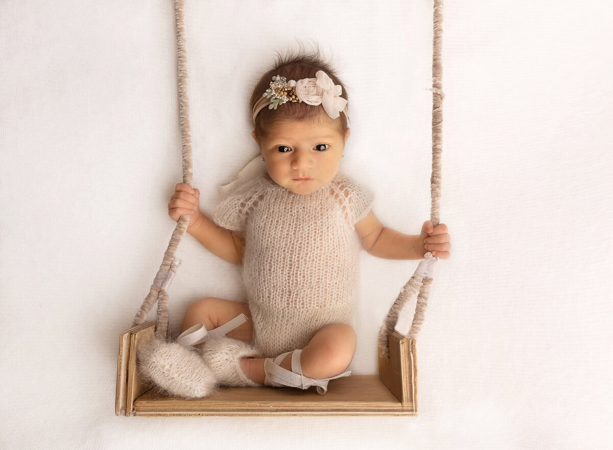 Baby girl in a beige knit romper and matching floral headband is laying on a blush fabric. There is a swing placed underneath her to give the illusion that she is sitting on a swing. Captured by top Brooklyn newborn photographer Chaya Bornstein