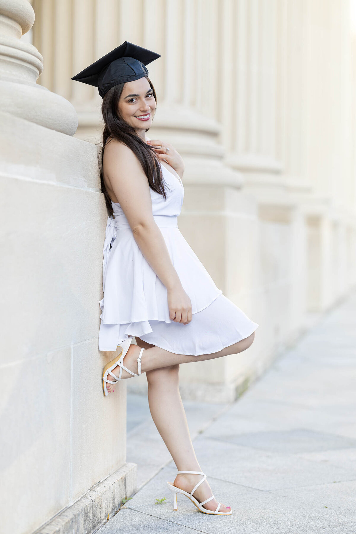 Graduate girl wearing a white dress leans against a column wall during her photo session in Connecticut