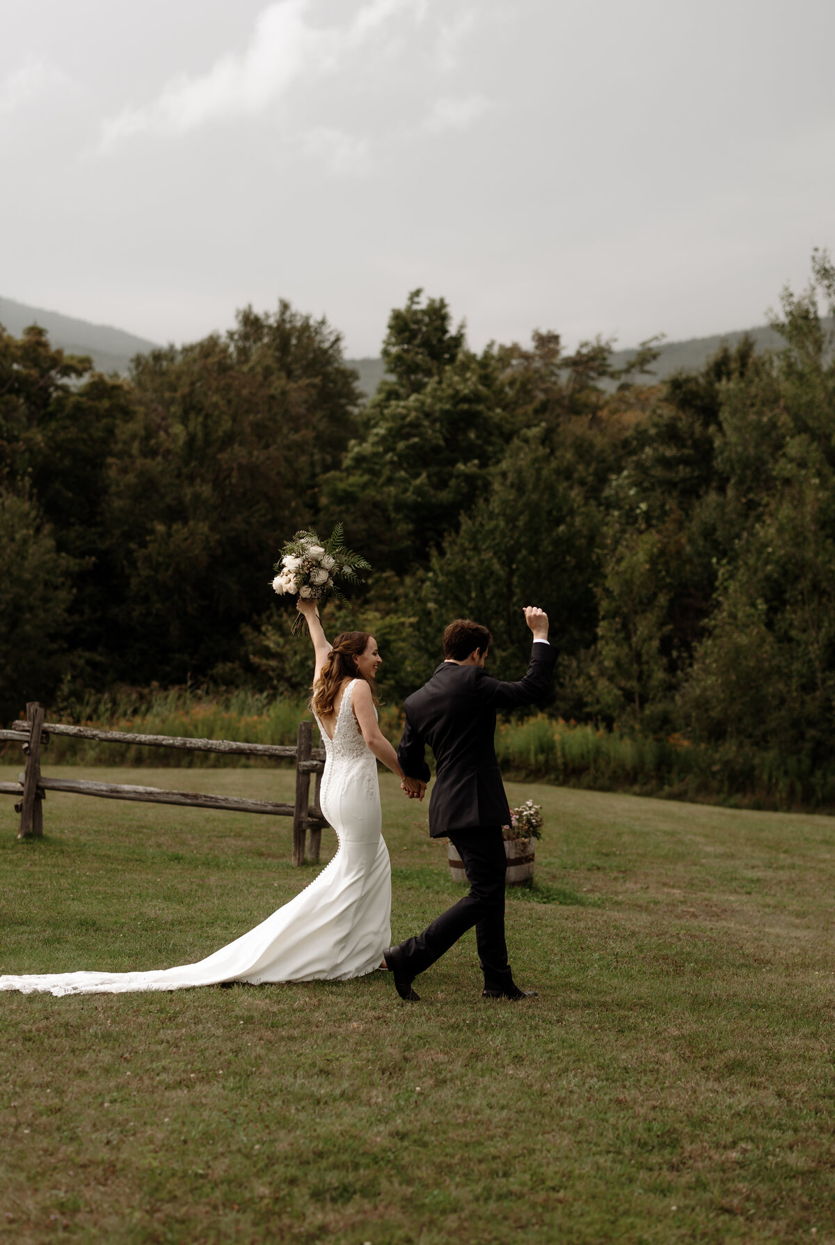 A bride and groom walk hand in hand across a grassy field, captured beautifully by a New England wedding photographer. The bride, in a white gown, raises her bouquet triumphantly as the groom, in a dark suit, holds his arm up. Trees and mountains frame this perfect moment under a cloudy sky.