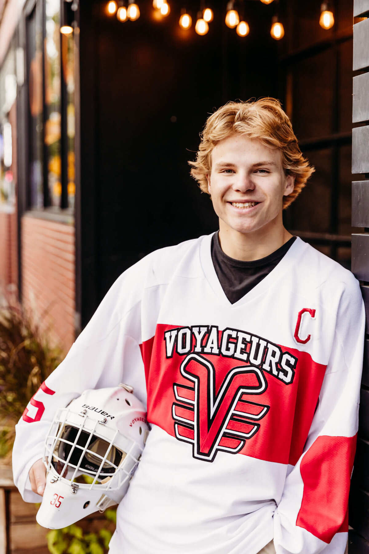 high school senior boy in a white hockey jersey leaning against a black wall and holding his hockey helmet while smiling 