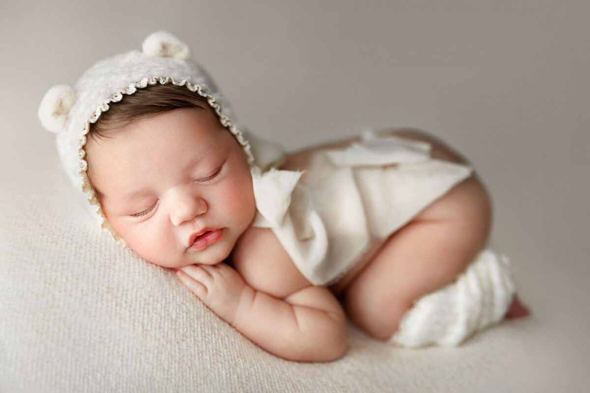 newborn baby in a bear   bonnet and leg warmers on a white backdrop at her newborn session