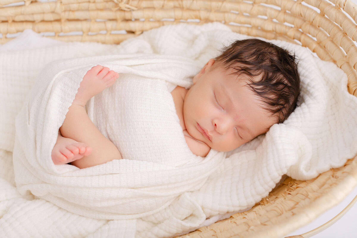 Newborn baby swaddled in white while laying in a Moses basket. Baby is sleeping peacefully.