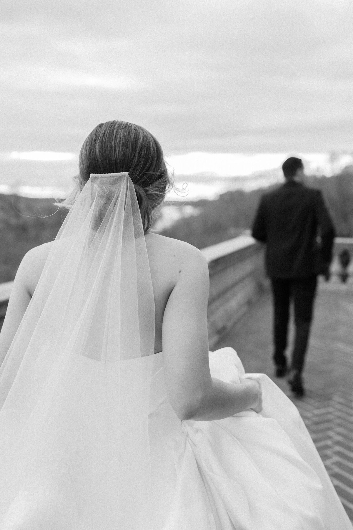 A timeless black-and-white photograph of a bride walking towards her groom at The Estate at River Run. Captured from behind, her delicate veil and elegant gown flow as she lifts her skirt slightly, following him along the brick terrace. The groom strides ahead, silhouetted against the soft, distant horizon. This moment, filled with quiet anticipation and romance, is beautifully documented by Caitlin Wilcox Photography, a Richmond, VA film wedding photographer.