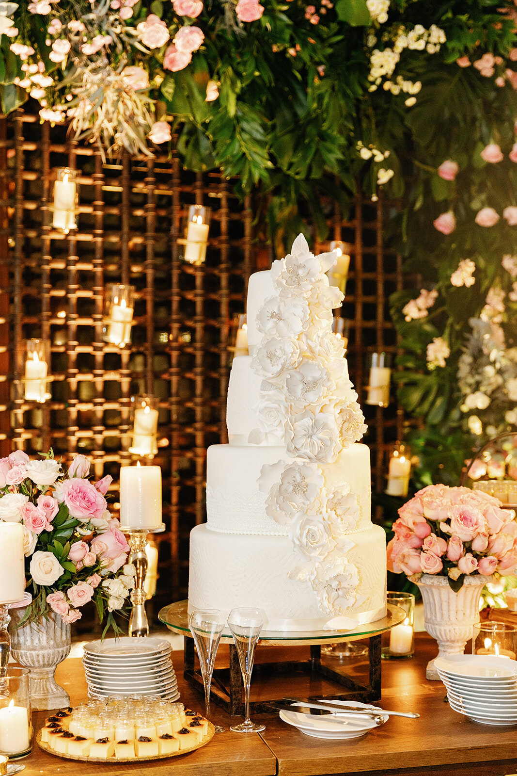 A breathtaking five-tier wedding cake decorated with intricate sugar flowers, set against a candlelit greenery backdrop.