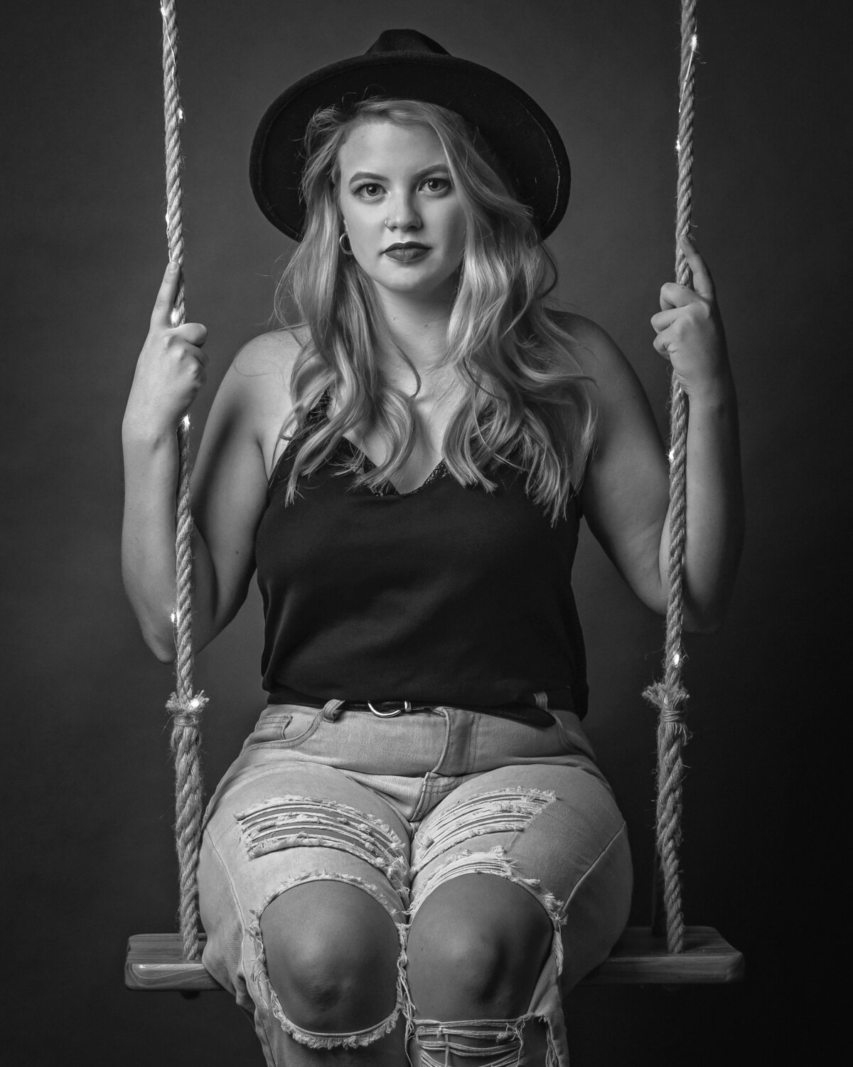 lady in ripped jeans, black hat, and black top seated upright on a wooden swing