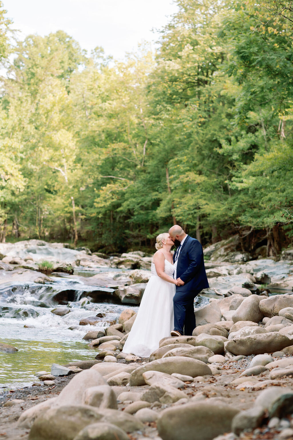 Bride and groom kissing on the rocky shoreline of the river at Greenbrier during their eloping to Gatlinburg celebration, with cascading water and dense forest in the background.
