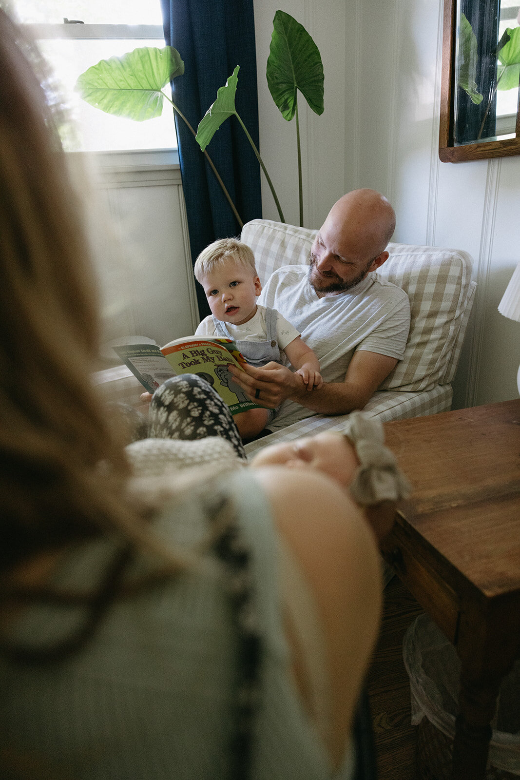 Little boy and his dad sit on a comfortable chair reading a children's book. 