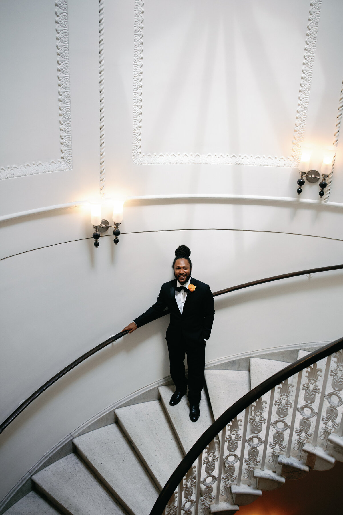Groom in classic black tuxedo standing on sweeping staircase at Hotel Monaco DC for wedding portraits.
