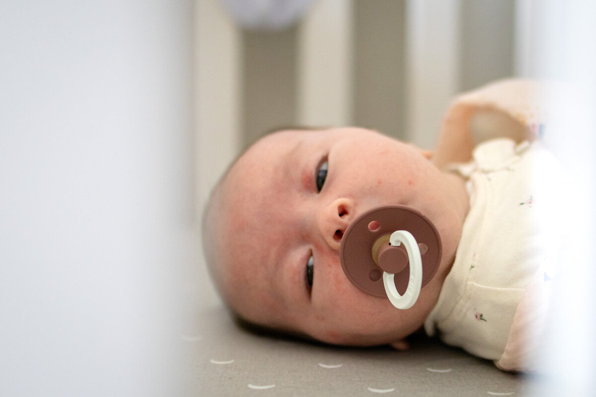 Newborn baby with pacifier lying in crib, viewed softly through crib slats – photographed by San Francisco newborn photographer.