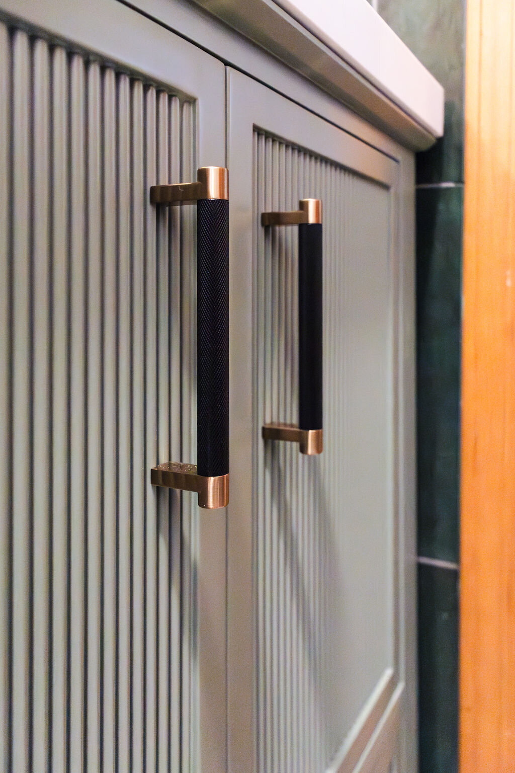 A close-up of a light sage green bathroom vanity cabinet with vertical ribbed detailing on the doors. Two modern, elongated cabinet pulls with a textured black center and brushed gold ends are affixed to the doors. The edge of a white countertop is visible at the top, and a section of dark green vertical tiled wall is on the right. The lighting is bright and highlights the texture of the cabinet doors and hardware.