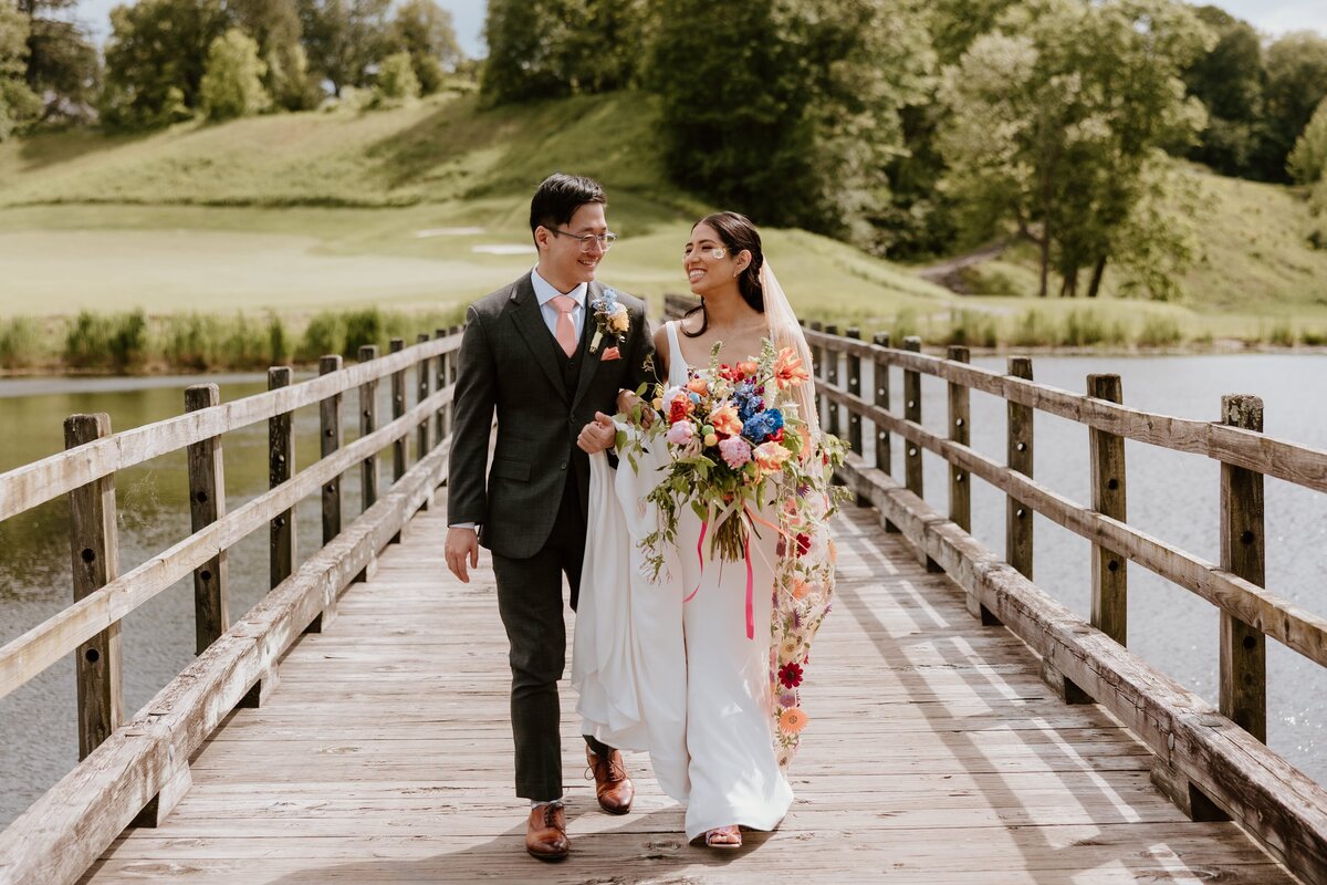 Bride and groom walk hand-in-hand across a wooden bridge at Hollow Brook Golf Club, smiling at each other under golden light with a vibrant, colorful bouquet.