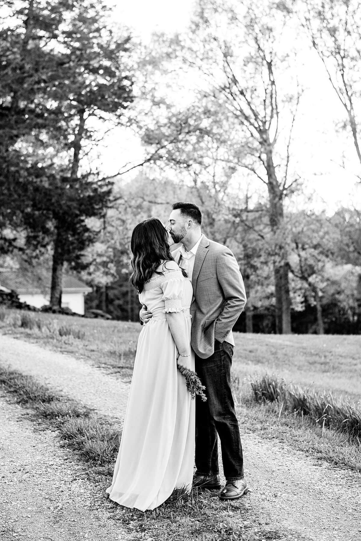 A black and white image of the bride and groom kissing on a gravel pathway next to a field with trees in the background. The bride is wearing a boho maxi dress with  short puffed sleeves . She holds her small bouquet down by her side. The groom is wearing a light color sports jacket and dark pants with a white shirt. He has one arm around the bride's waist and one hand in his pocket.