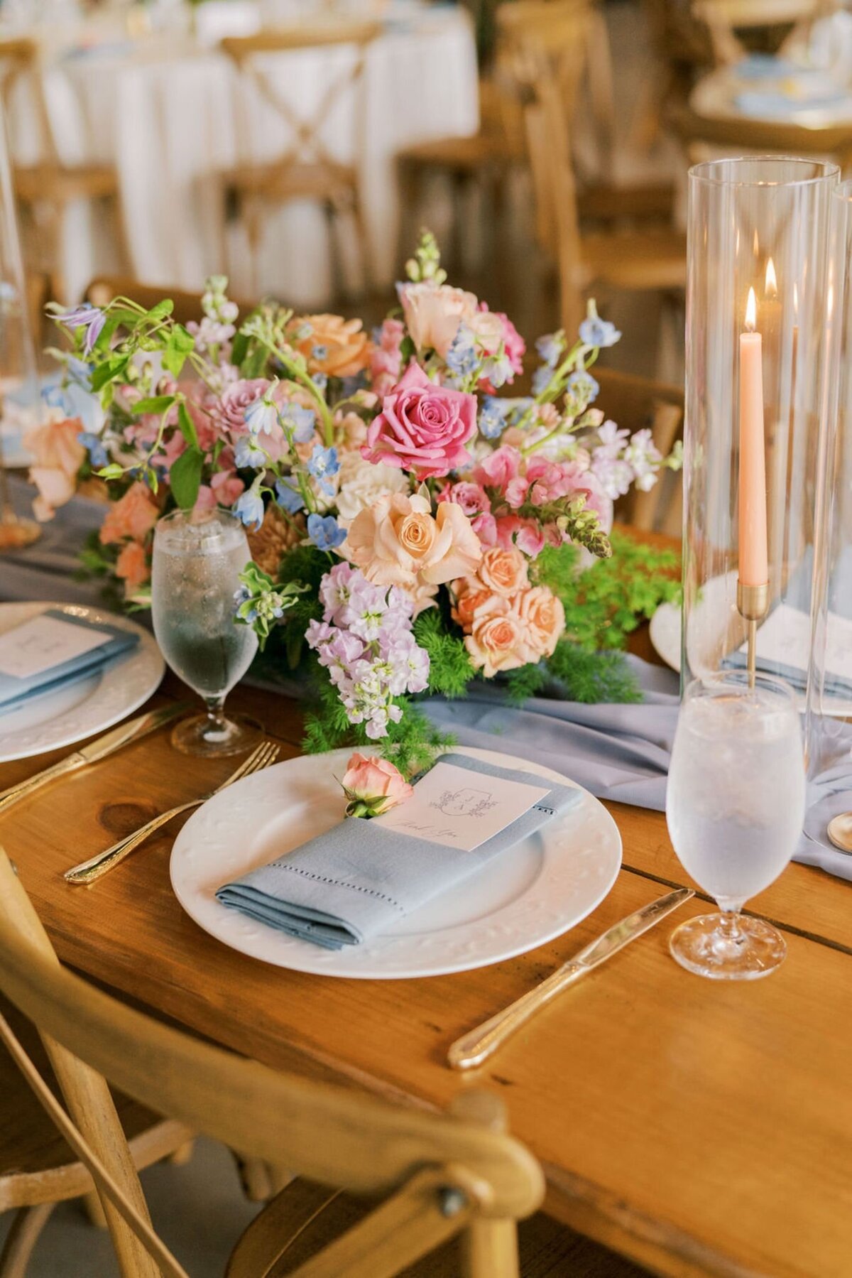 Soft pastel floral centerpiece with roses, delphiniums, and greenery on wooden reception table with blue linens.