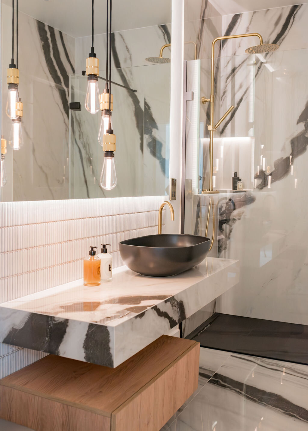 Modern bathroom with marble walls and countertops featuring black veining. A black vessel sink sits on a floating countertop with wooden storage below. Pendant lights hang above the sink, and a glass-enclosed shower with gold fixtures is visible in the background.