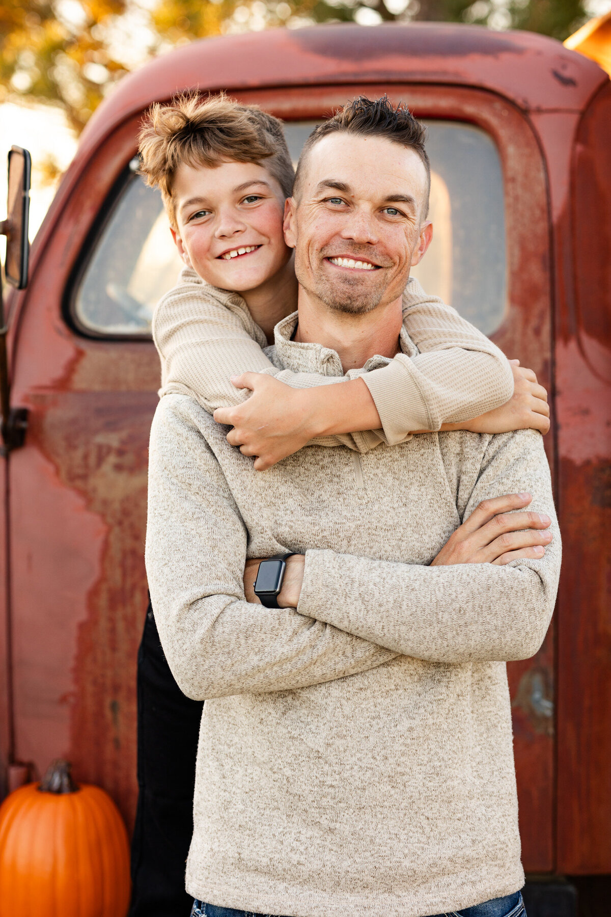Preteen boy stands behind his dad and hugs his dad around the neck as they both smile at the camera.