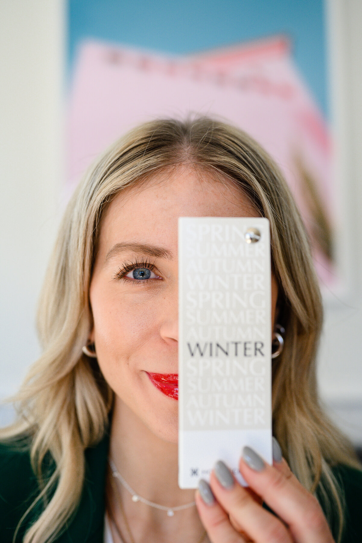 a personal brand photo of a close up of a woman with bright blue eyes and shiny red lipstick holding a card that says winter in front of one eye
