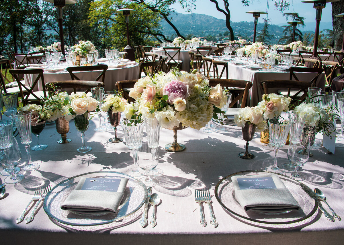 Tables are set up for a wedding outdoors with white and pink flowers on a white linen tablecloth.