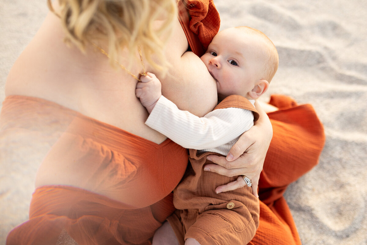 Mom breast-feeding infant boy in the sand while wearing a rust colored dress from Eden art dresses. 